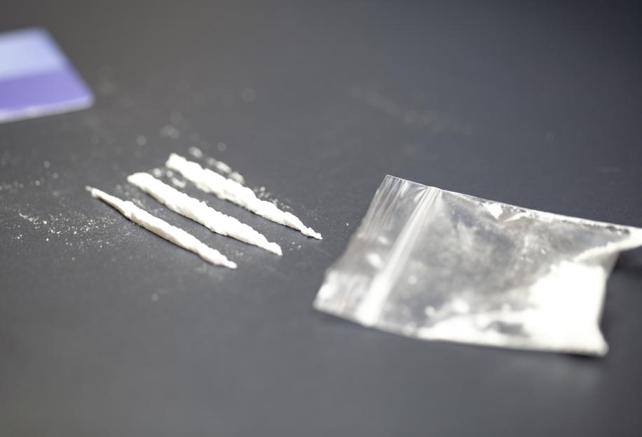 Four lines of cocaine next to a plastic bag of cocaine powder on a dark surface.