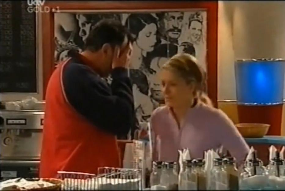 A man in a red and blue shirt covers his face in distress while talking to a blonde woman in a cafe.