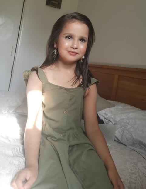 A girl with long dark hair, wearing a green dress and earrings, sitting on a bed.