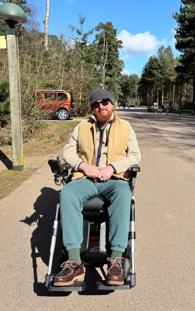 Liam Jefferies, a man with a red beard, wearing a beige vest, gray beanie, and sunglasses, sits in a power wheelchair on a paved path surrounded by pine trees.