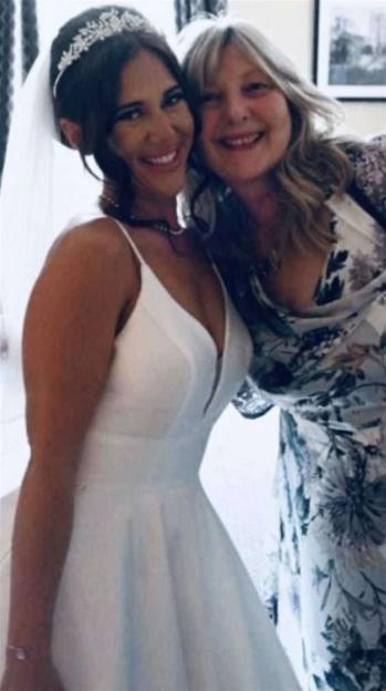 A smiling bride in a white dress and tiara poses next to an older woman with blonde hair wearing a floral dress.