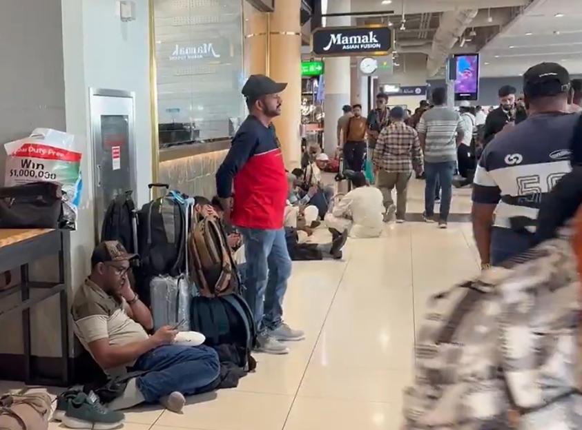 Passengers waiting in a crowded airport lounge, some sitting on the floor with their luggage.