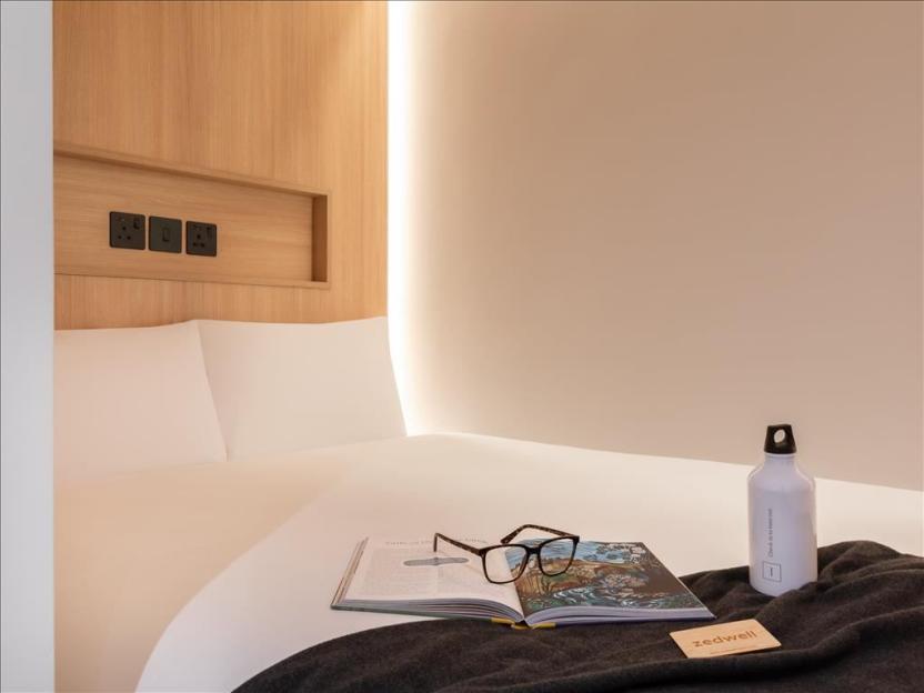 A Zedwell Park Lane hotel room featuring a bed, book, glasses, and water bottle.