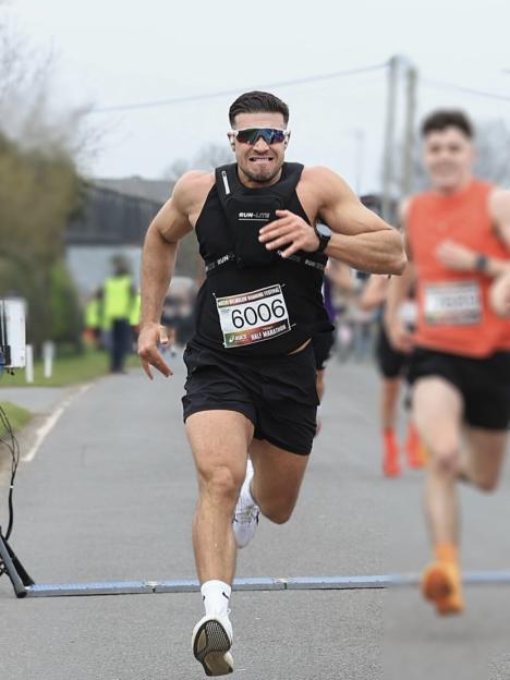 Tommy Fury sprinting across a marathon finish line, wearing a black running vest and bib number 6006.