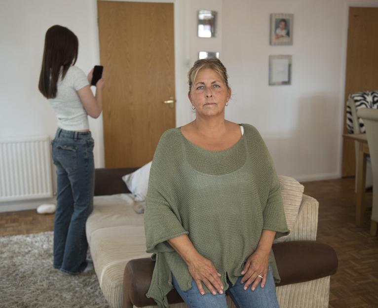 A woman sits on a sofa, looking at the camera, while her daughter stands behind her, looking at her phone.