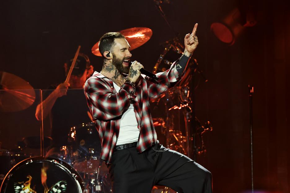 Adam Levine of Maroon 5 performs on stage, holding a microphone in one hand and pointing his finger up with the other.