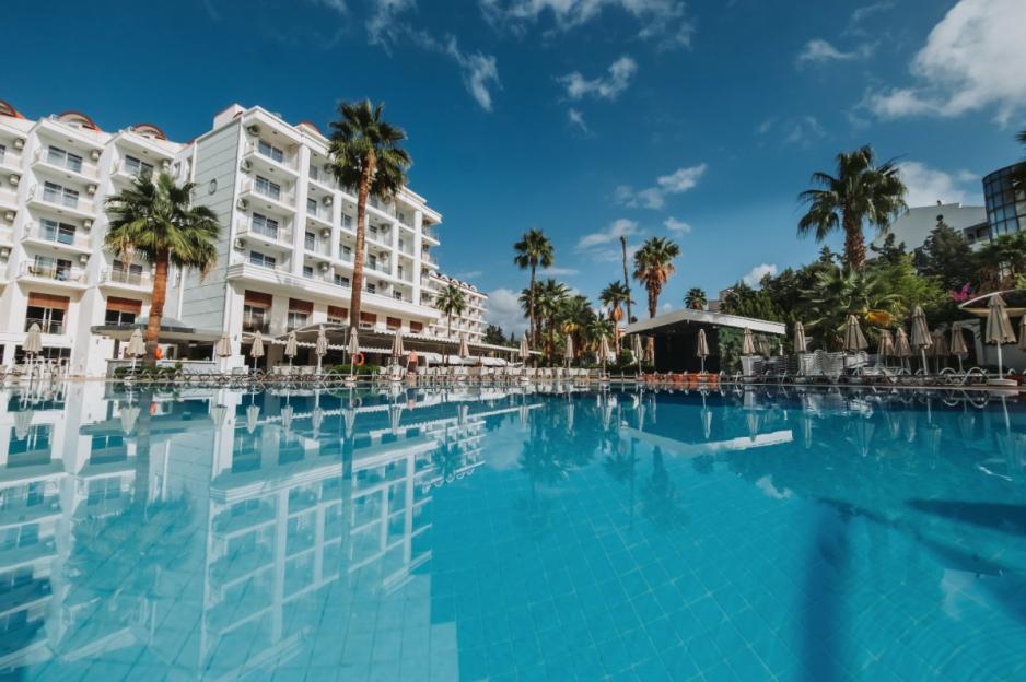 A hotel building, palm trees, and lounge chairs reflected in a swimming pool.