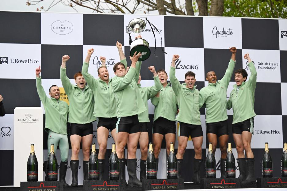 The victorious Cambridge men's rowing team celebrates their win in the Boat Race, holding up their trophy and bottles of sparkling wine.