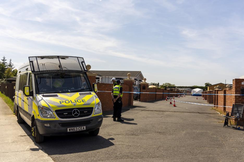 A police van at a crime scene with caution tape, cones, and a white tent in the background.