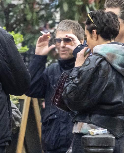 Liam Gallagher smoking a cigarette and adjusting his sunglasses while standing next to Debbie Gwyther.