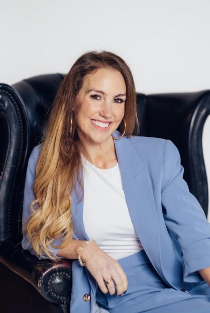 A smiling woman in a blue pantsuit sits in a black leather armchair.