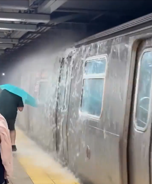 A person with a blue umbrella stands on a subway platform as heavy water cascades down the side of a parked subway train during flash flooding.