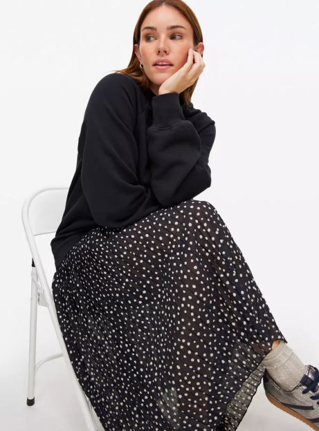 A woman wearing a black sweatshirt and black polka-dot skirt sits on a white chair.