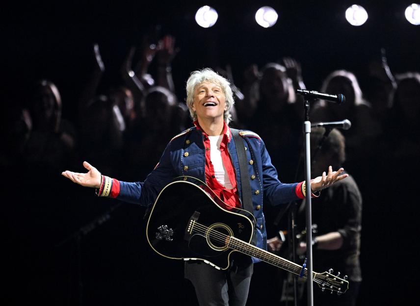 Jon Bon Jovi performing on stage at Wembley Stadium, wearing a blue jacket, white shirt, and black pants, with a black acoustic guitar.