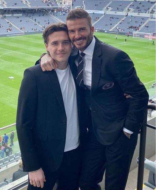 David Beckham with his arm around Brooklyn Beckham at a soccer stadium.