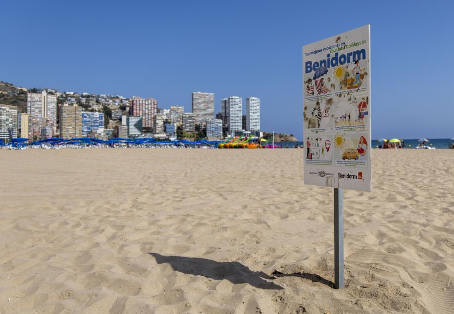 A sign on a sandy beach in Benidorm, Spain, provides tips for British holidaymakers, with city buildings and blue sky in the background.