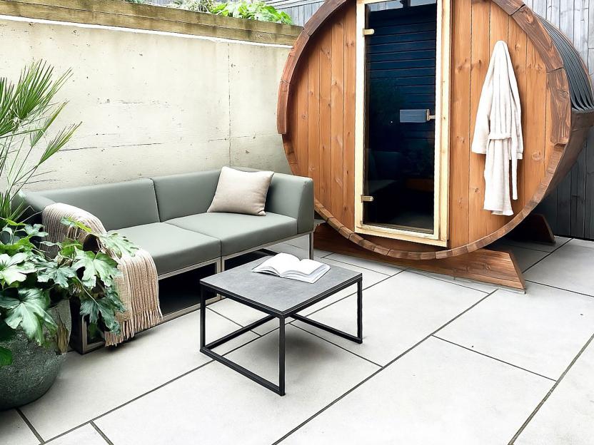 An outdoor patio with a gray couch, a coffee table, plants, and a wooden barrel sauna with a white bathrobe hanging on it.