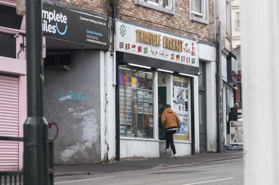 A man in a brown jacket enters a shop labeled "Triangle Market" and "Smoking Shop" with an adjacent dental practice.