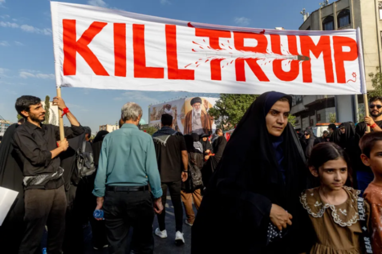 People marching with a banner that reads "KILL TRUMP" in red capital letters, with a bullet graphic creating a blood splat.