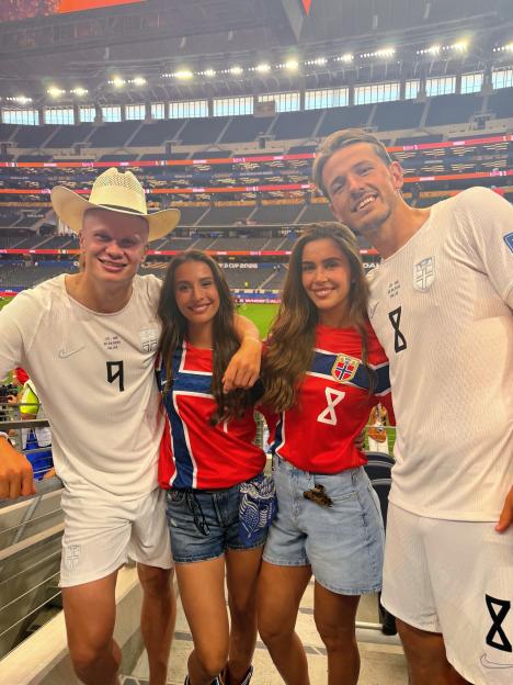 Four people, including Norwegian athletes Erling Haaland and Martin Ødegaard, pose for a photo in a stadium wearing Norway jerseys.