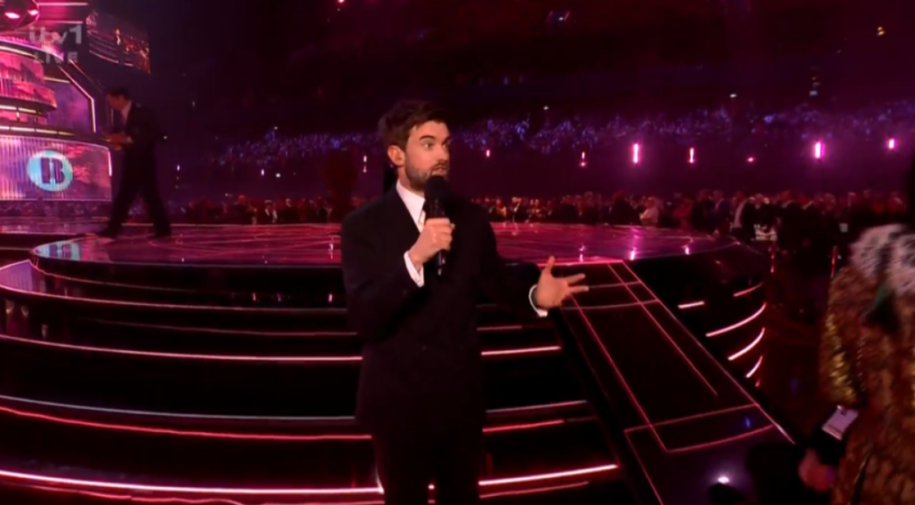 A man in a suit speaking into a microphone on a stage with glowing pink lights.