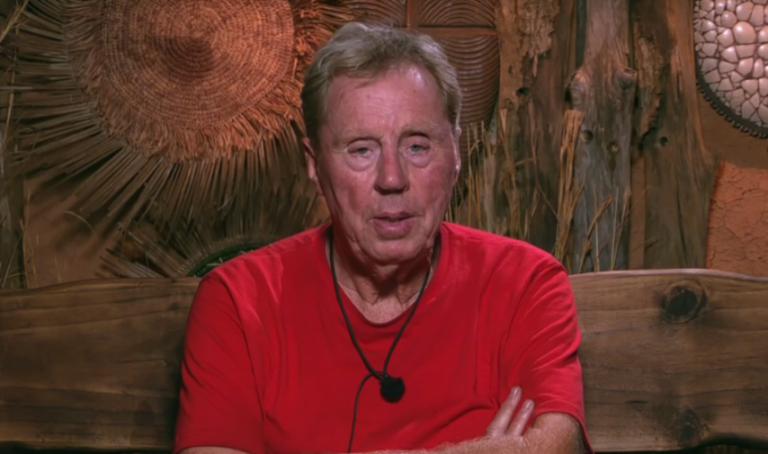 Harry Redknapp in a red shirt with his arms crossed, sitting on a wooden bench.