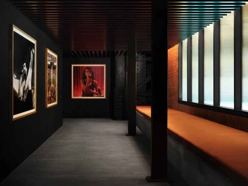 A long, dark corridor with framed photographs of musical artists on the left wall and a long orange bench under windows on the right.
