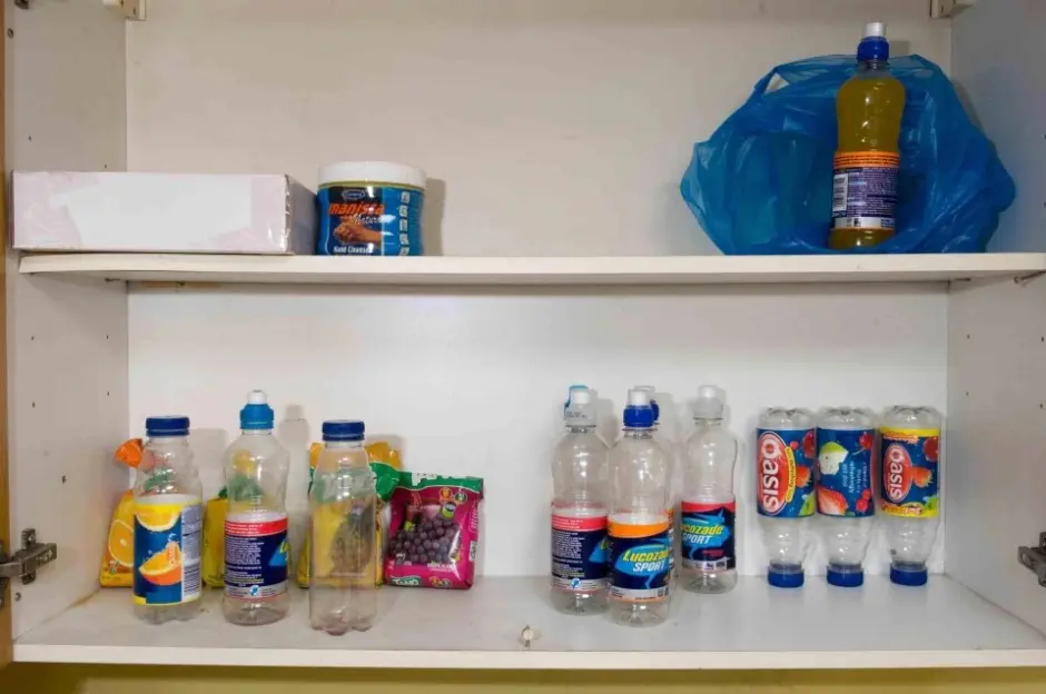 An open white cupboard with two shelves holding various plastic bottles, an unopened box, a blue tub, a plastic bag, and small snack bags.
