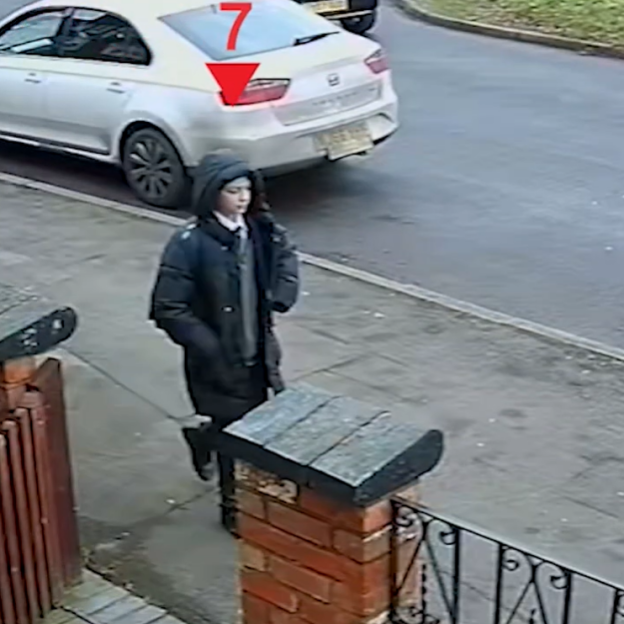 A person in a black coat and hoodie walks down a sidewalk next to a street with a parked white car.