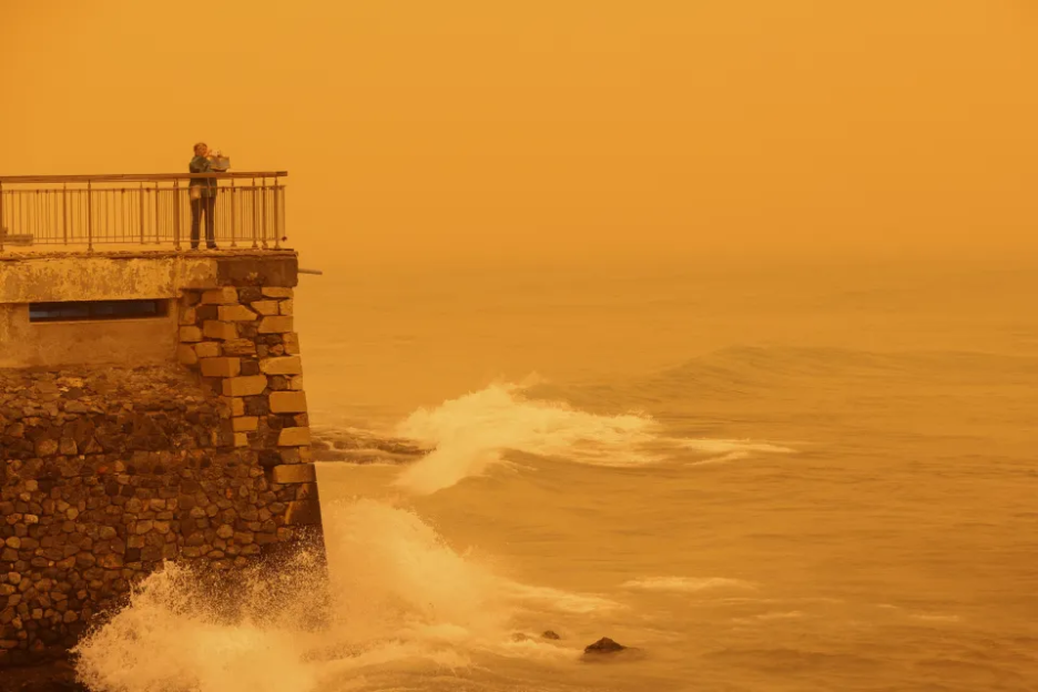 A person stands on a stone balcony overlooking an orange-tinted ocean with waves.