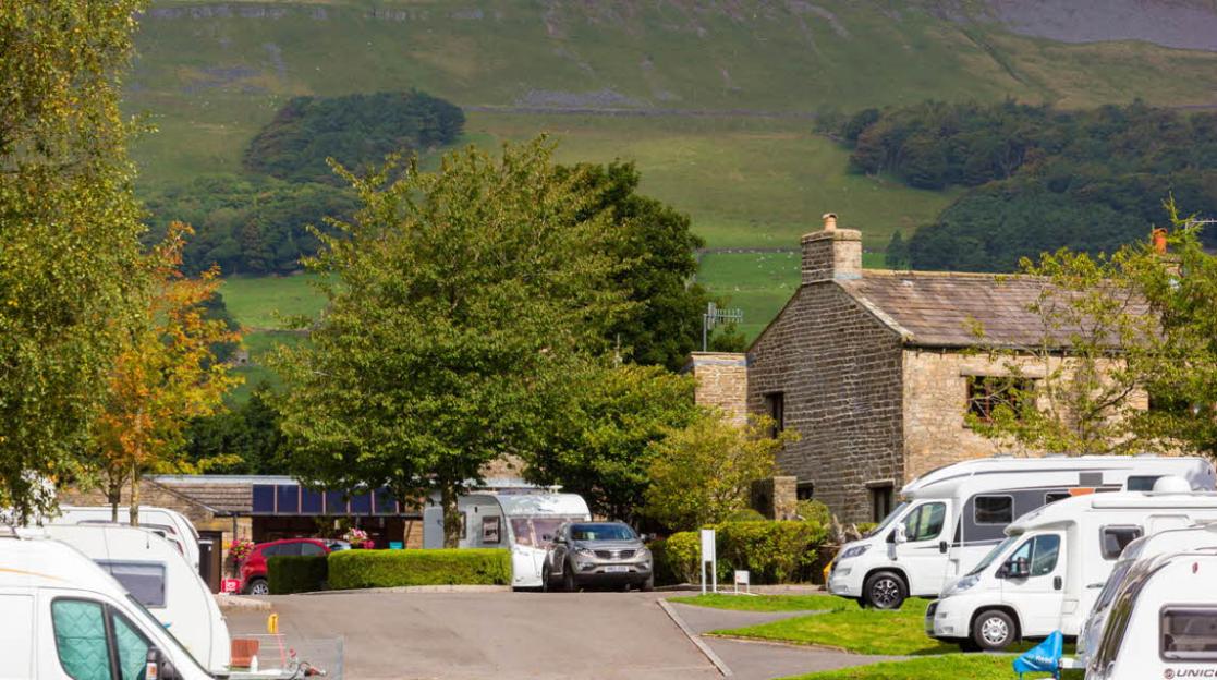 Hawes Club Campsite with multiple caravans and a brick building, set against a green hillside.