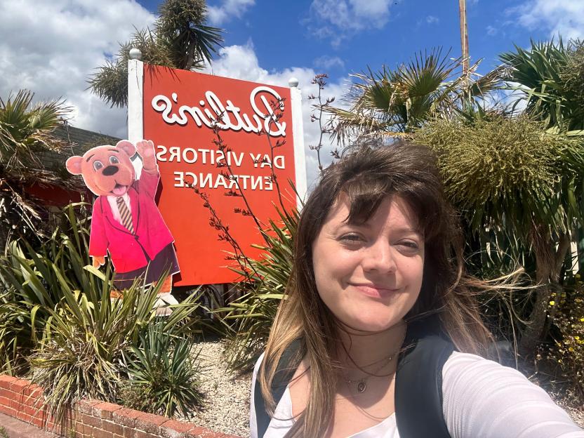 A woman smiles at the camera in front of a red "Butlin's Day Visitor Entrance" sign, with a cartoon bear cutout on the left.