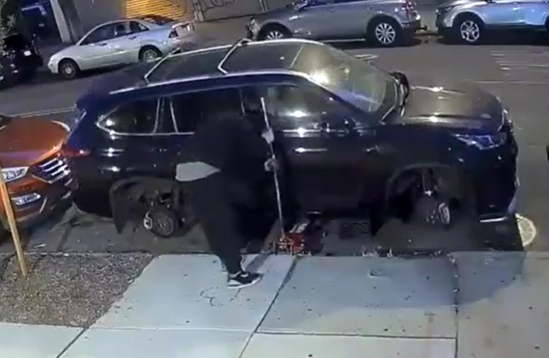 A person removing wheels from a dark SUV parked on the street.