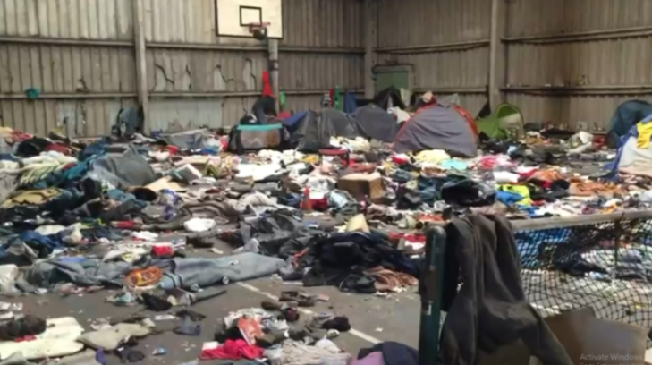 A large, cluttered warehouse or gym filled with scattered belongings, clothes, and several tents.