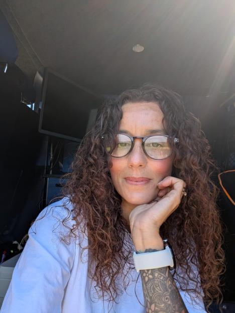 A woman with curly brown hair and glasses resting her chin on her hand, a tattoo on her forearm visible.