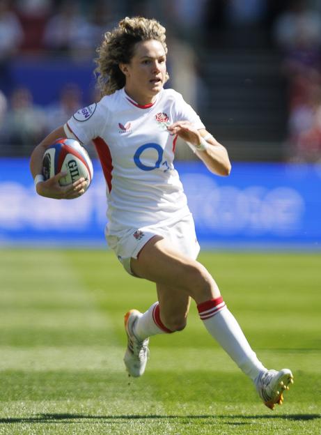 A female rugby player in a white uniform with red trim and an England rose emblem, running with a rugby ball.