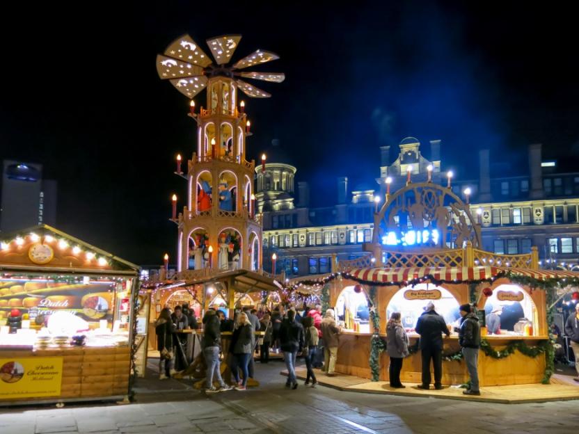 Manchester Christmas market by night with wooden stalls and a large lit Christmas pyramid.