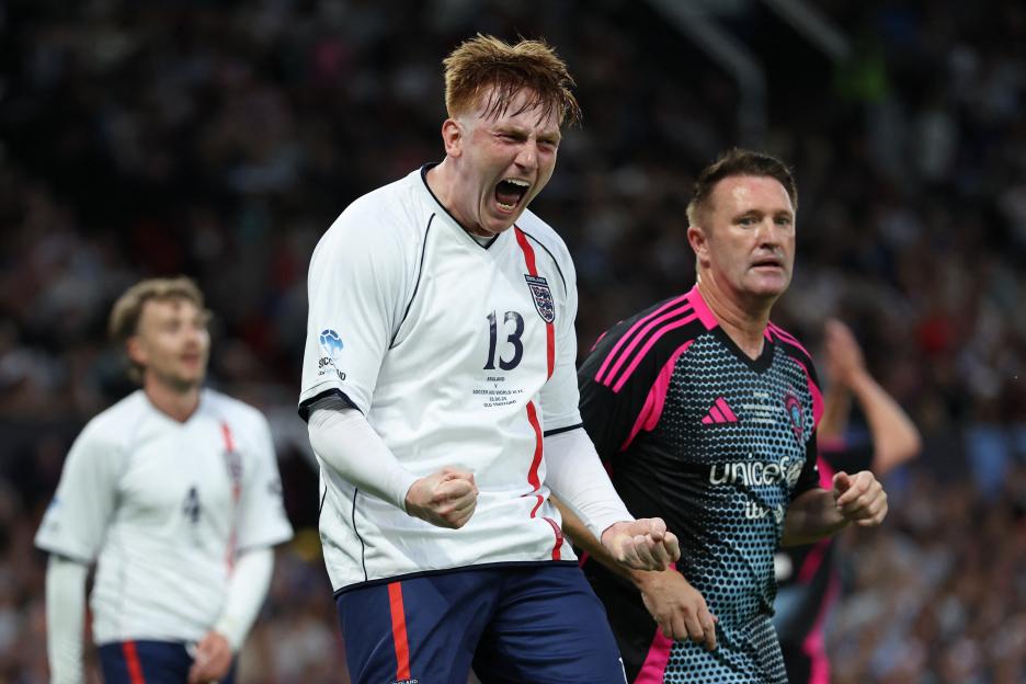 A soccer player in a white England jersey with the number 13 celebrating passionately, with another player in a black and pink UNICEF jersey watching him.