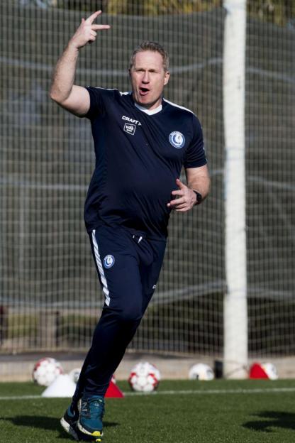 A man in a navy blue tracksuit making a "rock and roll" hand gesture while shouting on a soccer field.