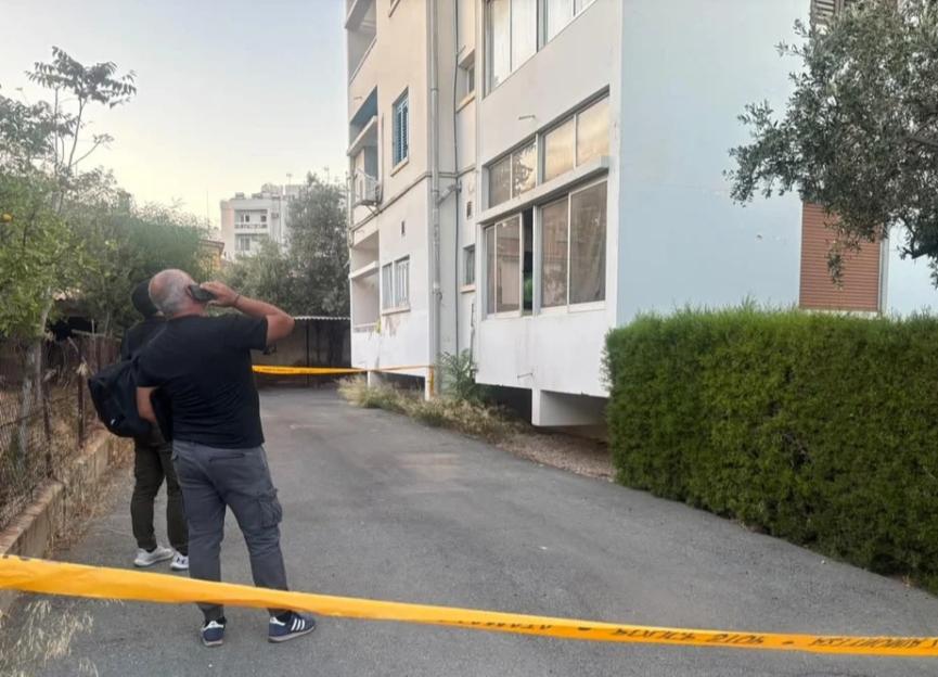 Two men stand behind yellow police tape in front of a residential building.