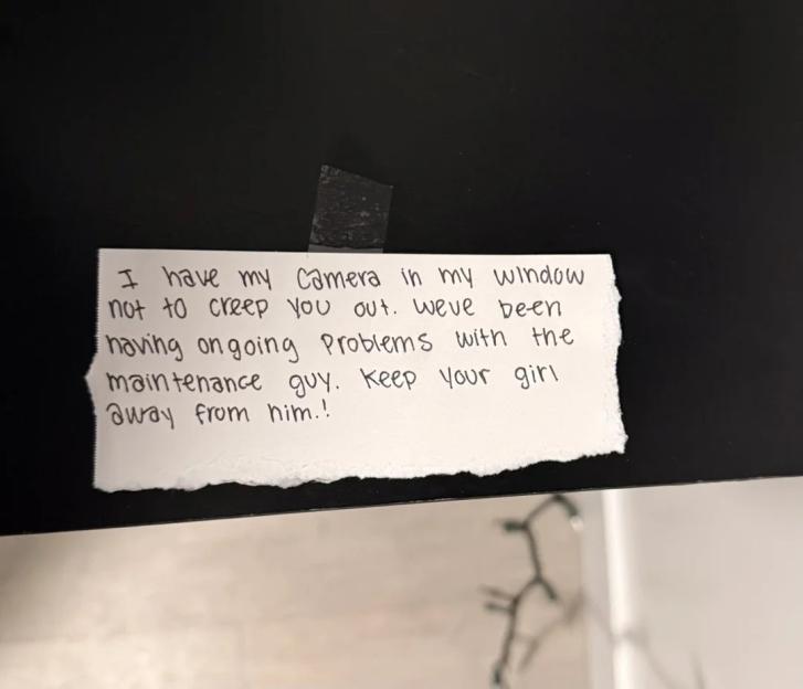 A handwritten note on torn white paper taped to a dark surface reads: "I have my Camera in my window not to creep you out. weve been having ongoing Problems with the maintenance guy. keep your girl away from him.!"