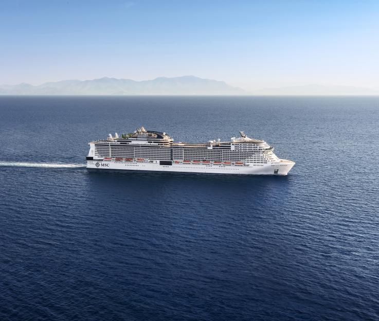Aerial view of the MSC Virtuosa cruise ship sailing on blue waters with mountains in the background.