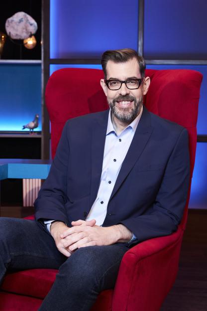 Richard Osman smiles while sitting in a red armchair.