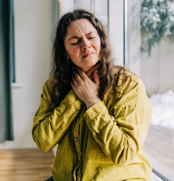 A woman with long curly hair holds her throat with both hands, eyes closed in pain, likely suffering from a sore throat.