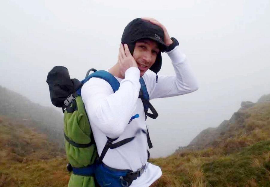 Kristian Hudson wearing a black hat and white long-sleeved shirt with a green and blue backpack, in a foggy outdoor setting.
