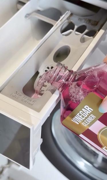 A person pouring pink liquid labeled "Vinegar Cleaner" into the "Softener Max 100ml" compartment of a washing machine drawer.