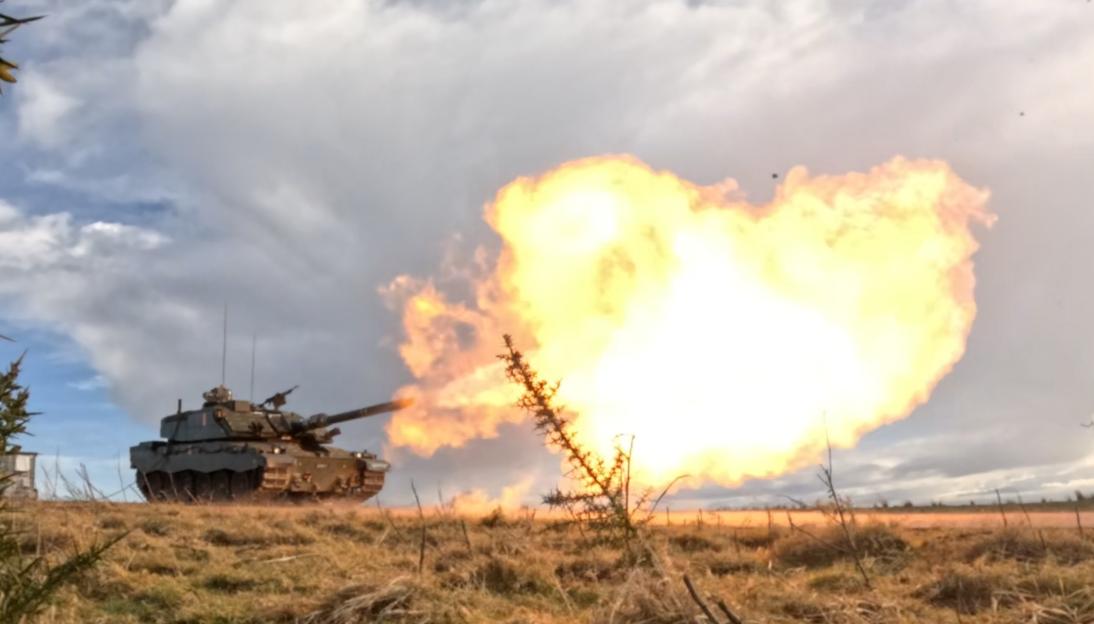 A tank firing, with a large explosion of fire and smoke erupting from its barrel, against a cloudy sky.