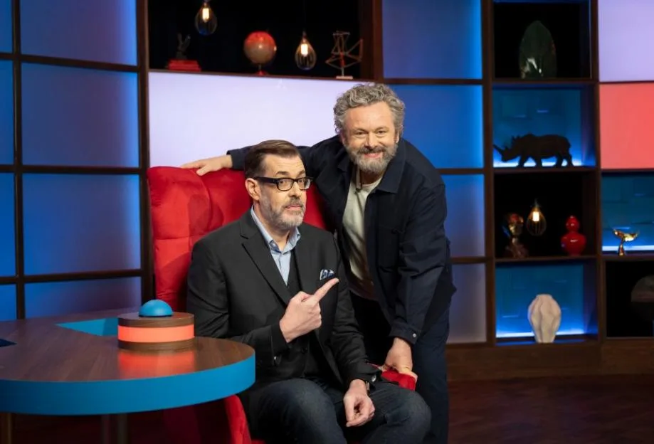 Richard Osman sits in a red armchair, pointing to his right, while Michael Sheen leans over him, smiling at the camera, on a television game show set.