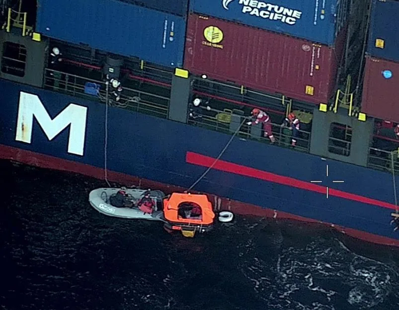 An aerial view shows a cargo ship conducting a rescue operation involving an inflatable boat and an orange life raft in the water.