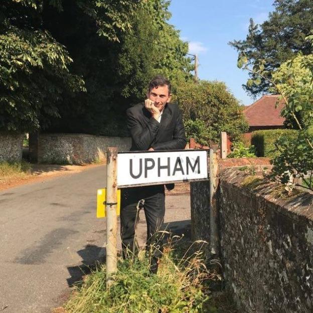 A man in a suit poses with a sign that reads 'UPHAM.'